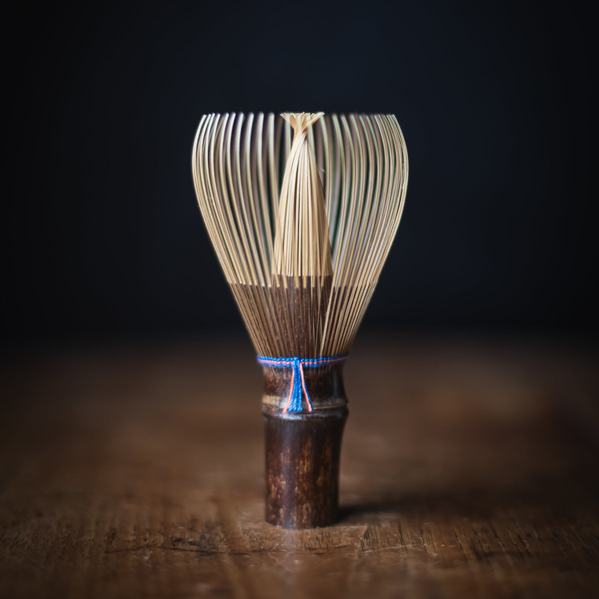 A traditional Japanese bamboo whisk (chashaku) with a dark brown wooden handle and a blue decorative band, standing on a rustic wooden surface against a dark background.
