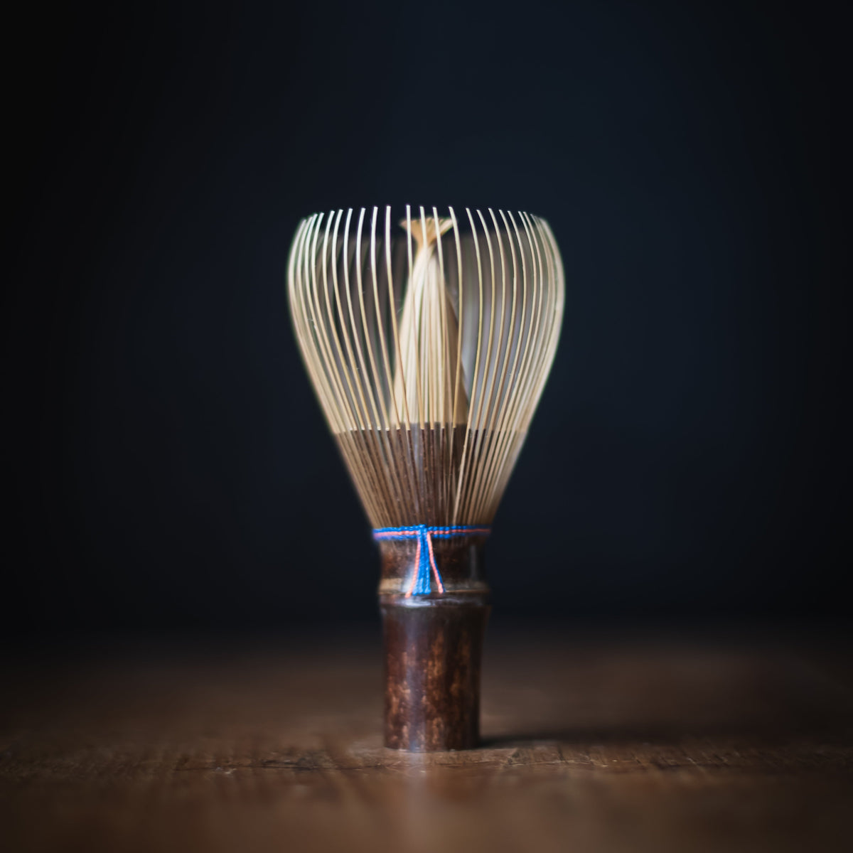 A traditional Japanese bamboo whisk (chashaku) with a dark brown wooden handle and a blue cord accent, standing on a wooden surface against a dark background.