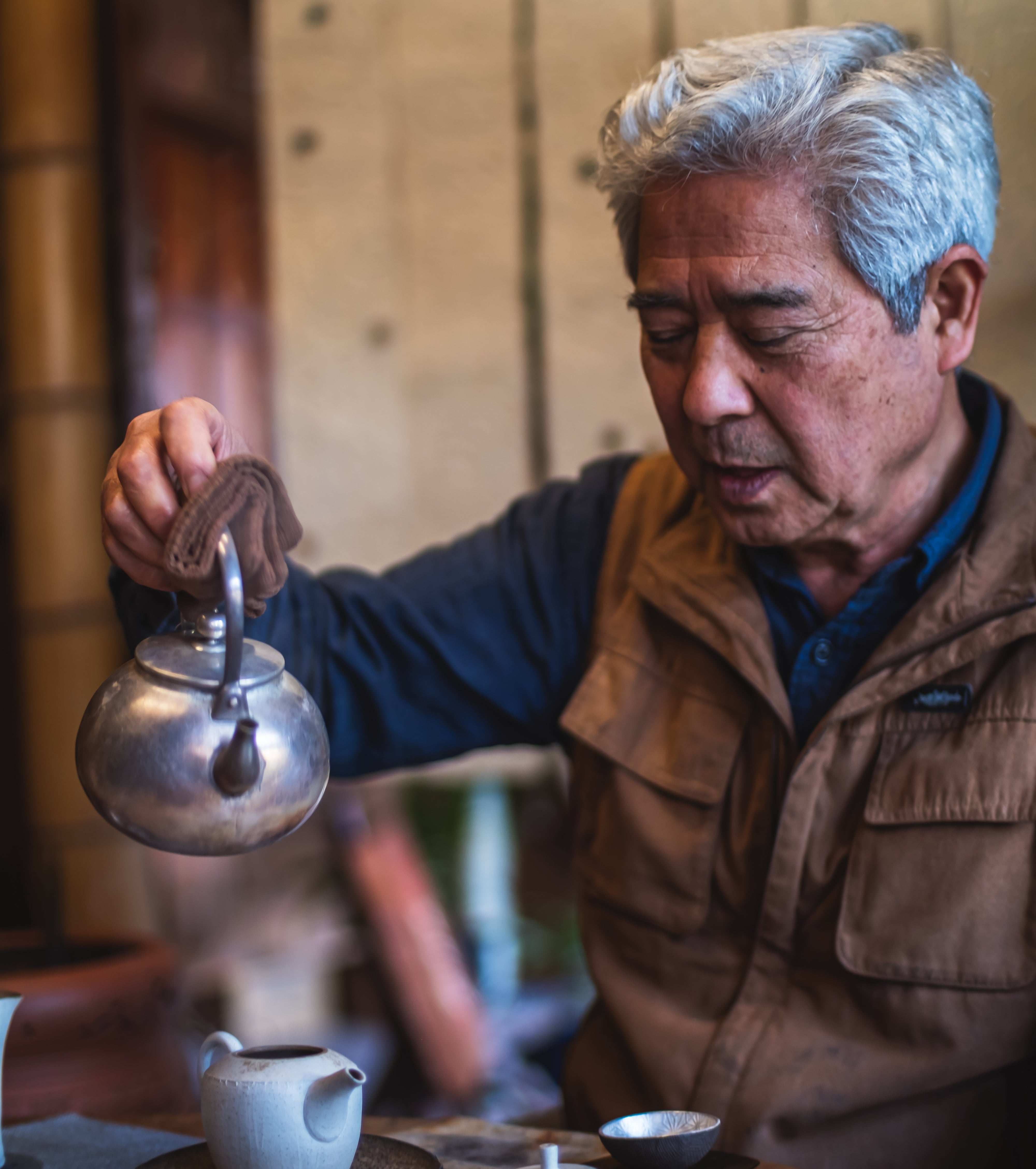 Ceramicist Seiji Ito pouring tea from a silver kettle into a teapot on a wooden table in his owm tea room.