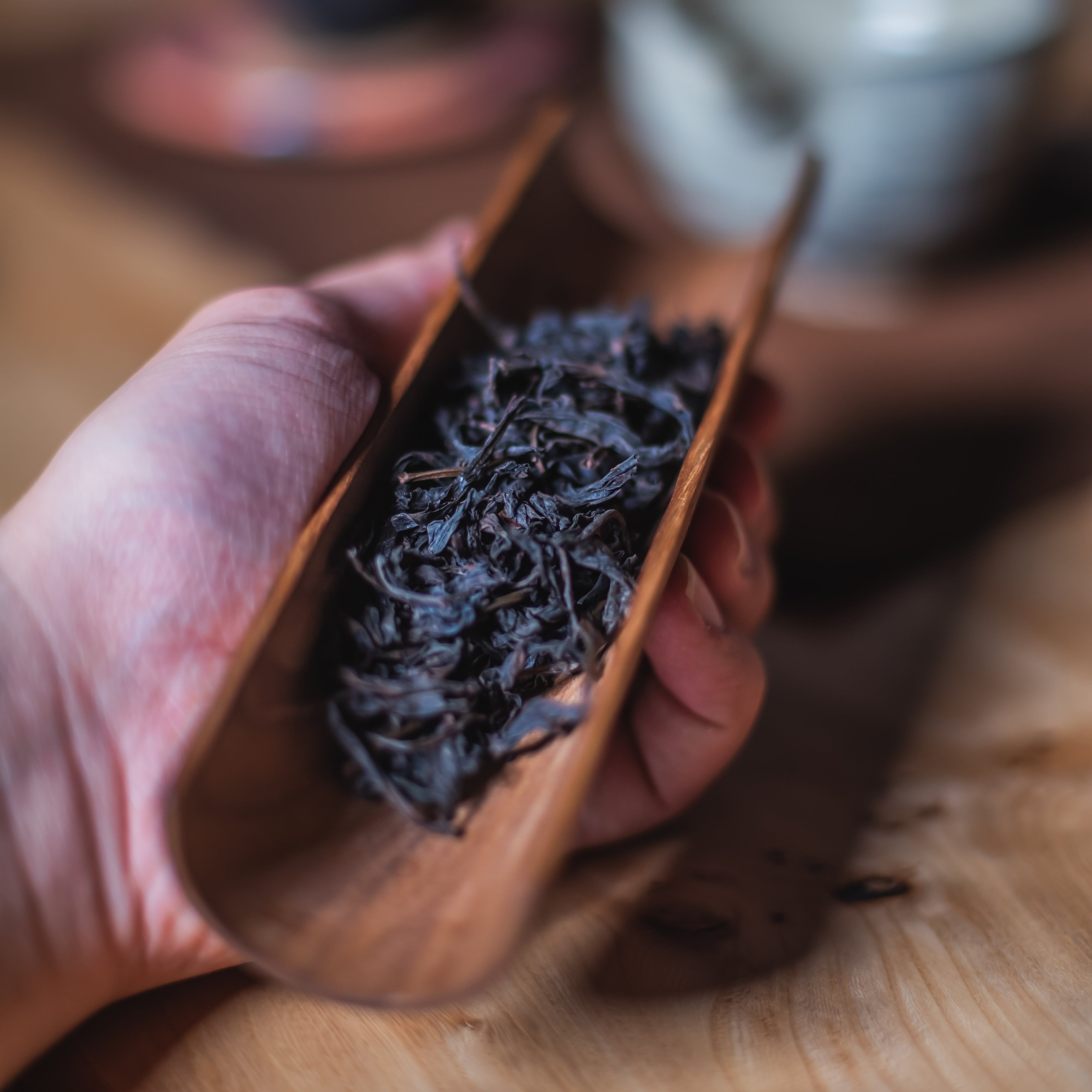 Hand holding a wooden scoop filled with dark dried leaves on a wooden surface.