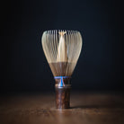 A traditional Japanese bamboo whisk (chashaku) with a dark brown wooden handle and a blue cord accent, standing on a wooden surface against a dark background.