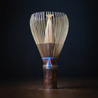 A traditional Japanese bamboo whisk (chashaku) with a dark brown wooden handle and a blue cord accent, standing upright on a dark wooden surface against a dark background.