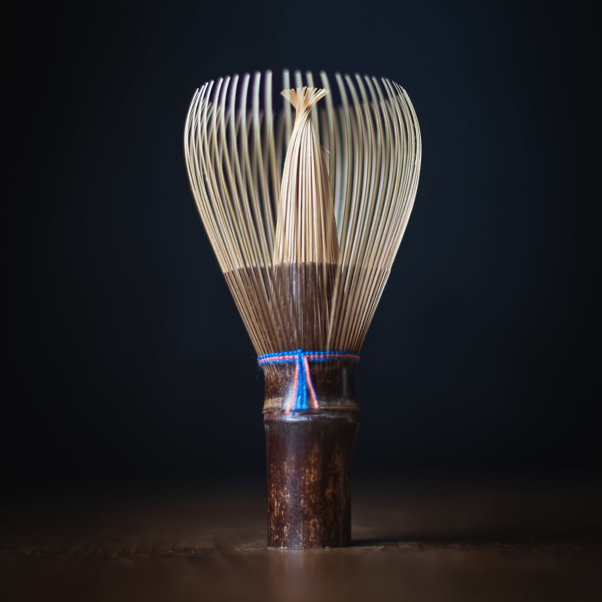 A traditional Japanese bamboo whisk (chashaku) with a dark brown wooden handle and a blue cord accent, standing upright on a dark wooden surface against a dark background.