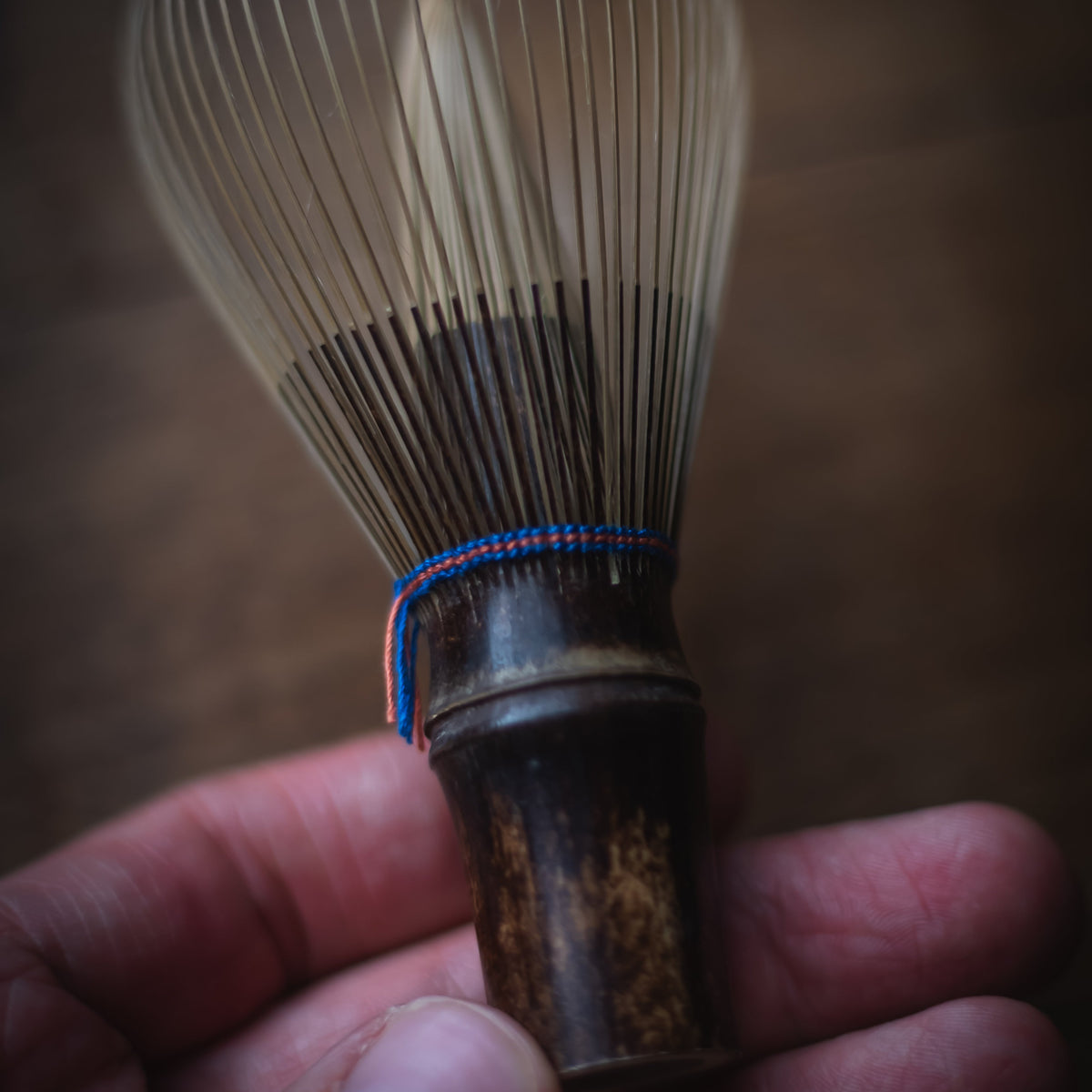 A close-up of a hand holding a traditional bamboo whisk with fine light brown bristles and a dark brown handle, tied with a small blue and red cord, against a dark blurred background.