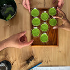 Overhead view of a wooden tray holding seven small clear cups filled with bright green matcha tea, with several hands reaching toward it on a light wood table. A dark green bowl, whisk, blue pencil, and printed sheet are also visible.
