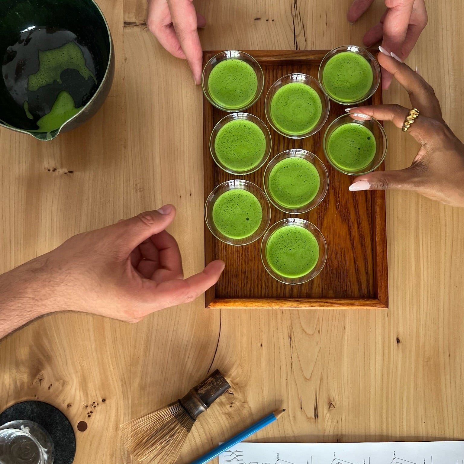 Overhead view of a wooden tray holding seven small clear cups filled with bright green matcha tea, with several hands reaching toward it on a light wood table. A dark green bowl, whisk, blue pencil, and printed sheet are also visible.